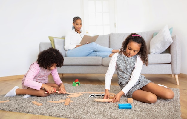 Czarne córki bawiące się zabawkową kolejką, podczas gdy matka czyta w pomieszczeniu