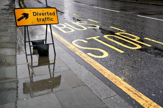 Czarno-żółty znak przekierowania ruchu na mokrym chodniku, obok drogi z napisem Bus Stop, światła odbijają się w pokrytym deszczem asfalcie