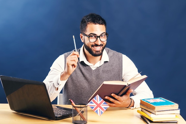 Czarnowłosy mężczyzna uczący się angielskiego ubrany w szarą kamizelkę swetrową i nieskazitelnie białą koszulę uśmiechający się do biurka z laptopem przed czarną ścianą