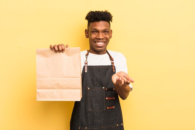 Czarny afro młody mężczyzna uśmiechający się radośnie z przyjaznym i oferującym koncepcję zabierania koncepcji pracownika żywnościowego