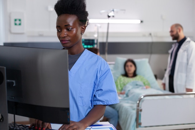 Czarny asystent medyczny wpisując leczenie choroby na komputerze, podczas gdy lekarze lekarze monitorują chorą kobietę analizując diagnozę. Pacjent leżący w łóżku oddziału szpitalnego podczas powrotu do zdrowia po konsultacji