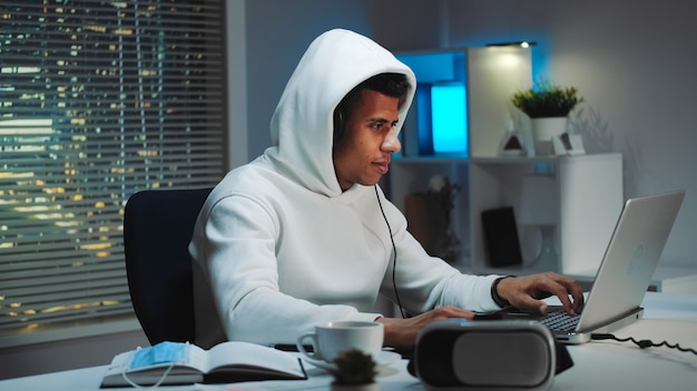 Czarny gracz w białej bluzie i słuchawkach grający wieczorem w gry na komputerze. Jest namiętny i uśmiechnięty. Wieżowce w tle.