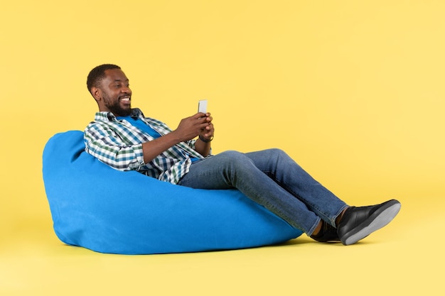 Czarny mężczyzna korzystający z telefonu komórkowego siedzącego na krześle beanbag w studio