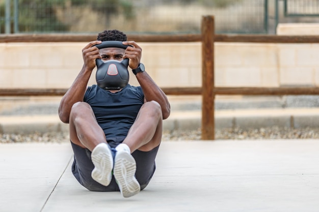 Czarny mężczyzna w odzieży aktywnej wykonujący ćwiczenie brzucha z kettlebellem podczas ćwiczeń na świeżym powietrzu