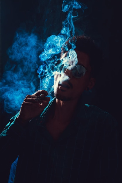 Czarny młody człowiek w okularach palący cygaro z mgłą na czarnym tle