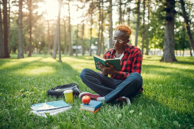 Czarny student w okularach, czytanie książki na trawie