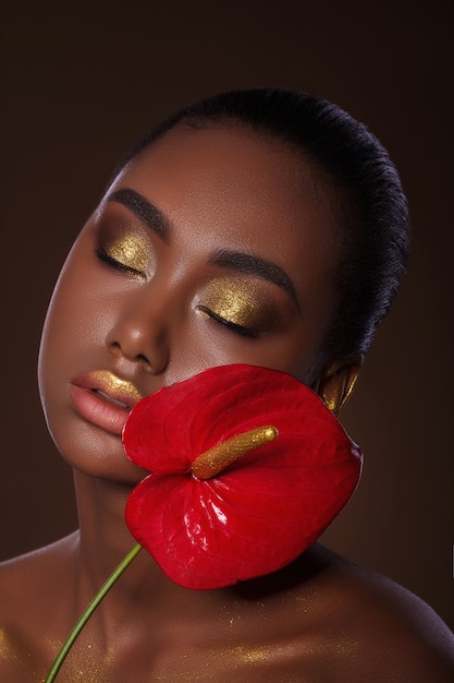 czarujący afrykański portret dziewczyny z czerwonym egzotycznym kwiatem. złoty makijaż. Piękna twarz.