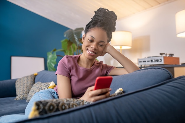 Czas dla siebie. Młoda dorosła piękna afroamerykańska kobieta w koszulce patrząca uśmiechnięta na smartfona siedzącego na kanapie