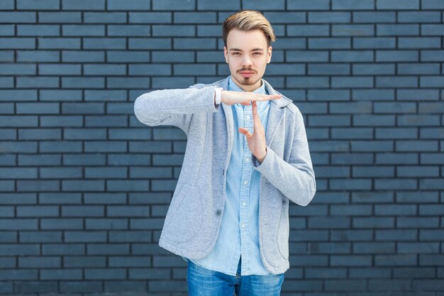 Zdjęcie czas portret poważne przystojny młody blondyn w stylu casual stojący z limitem czasu gest ręką i patrząc na kamery ze spokojną twarzą kryty studio strzał na tle ściany z cegły