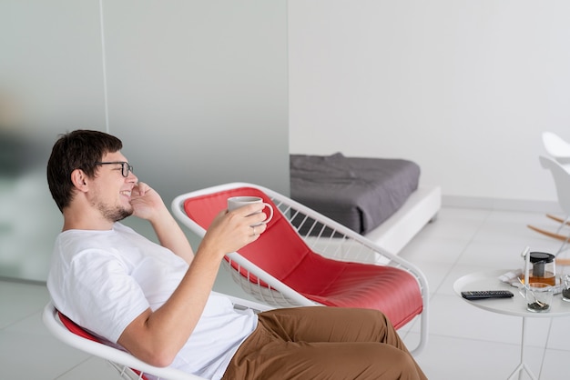 Czas wolny. Mężczyzna w białej koszuli siedzi na krześle oglądając telewizję, trzymając filiżankę herbaty i telefon
