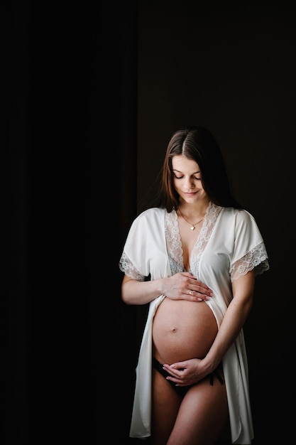 Czeka na dziecko Seksowna kobieta w ciąży stojąca przy oknie w domu i ręce obejmuje okrągły brzuch z bliska dziewięć miesięcy Baby Shower Koncepcja macierzyństwa Miejsce na tekst