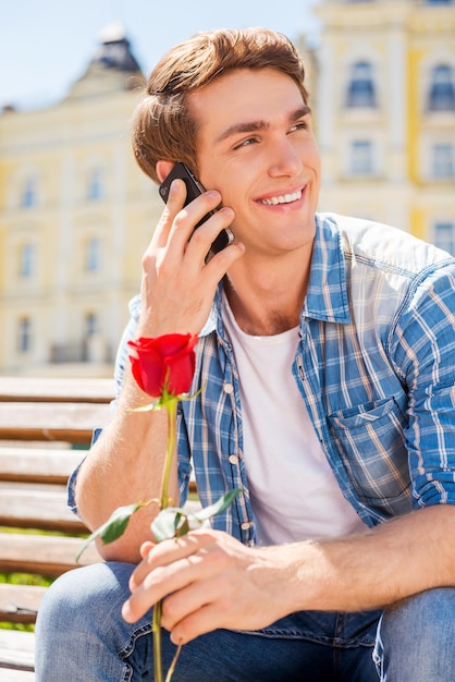 Czekam na Ciebie! Szczęśliwy młody człowiek trzyma pojedynczą różę i rozmawia przez telefon komórkowy, siedząc na ławce