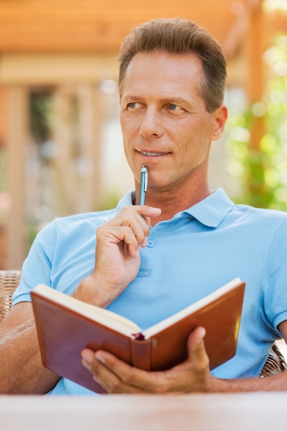 Czekam na inspirację. Rozważny dojrzały mężczyzna trzymający notatnik i dotykający podbródka długopisem podczas siedzenia na zewnątrz