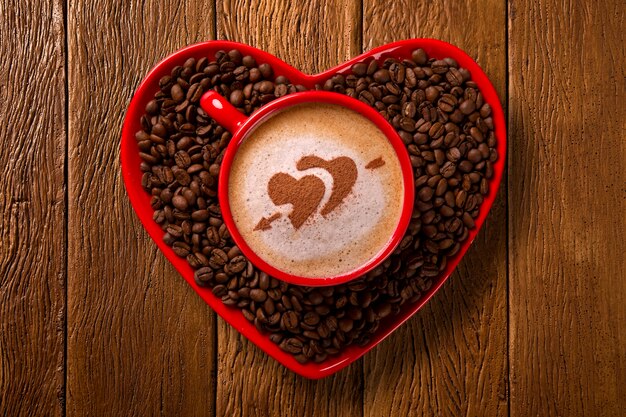 Czerwona filiżanka i spodek do kawy w kształcie serca z dekorowaną kawą na starym drewnie. Widok z góry. Kształt serca w kawie.