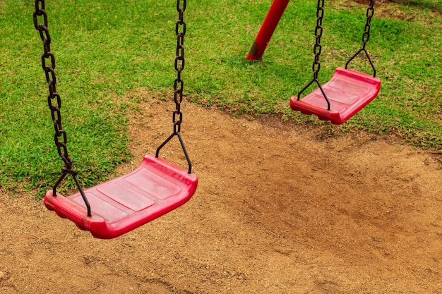 Czerwona huśtawka jest przymocowana do dwóch starych zardzewiałych łańcuchów na placu zabaw w parku, piła ziemię i trawę