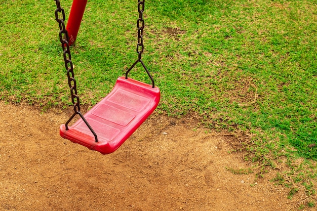 Czerwona huśtawka jest przymocowana do zardzewiałego, starego łańcucha na placu zabaw w parku, piła ziemię i trawę