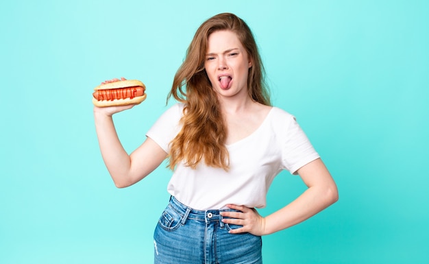 Czerwona ładna kobieta czuje się zdegustowana i zirytowana, wysuwa język i trzyma hot doga