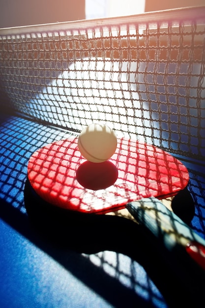 Czerwona rakieta do tenisa stołowego i biała piłka leżą na powierzchni stołu obok siatki sportowej