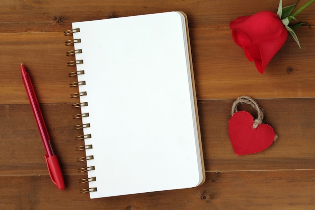 Czerwona róża, tkaniny serca kształt i pusta notatka książki papieru na tle drewna
