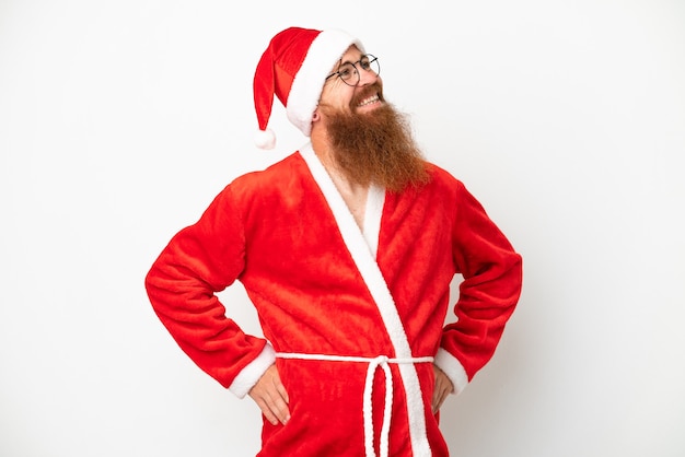 Czerwonawy mężczyzna przebrany za Świętego Mikołaja na białym tle, pozujący z rękami na biodrach i uśmiechnięty