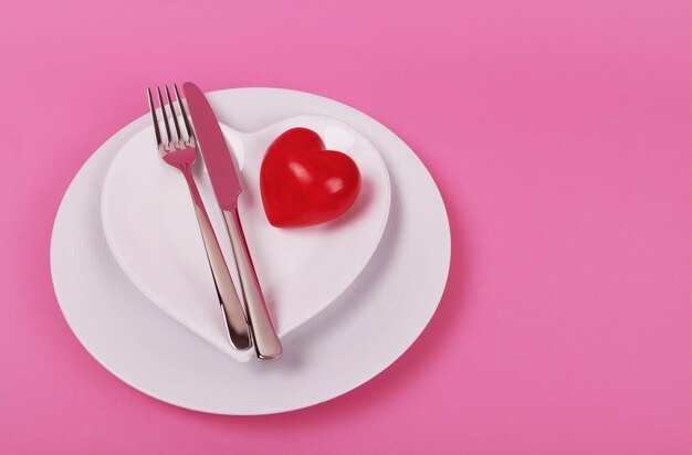 Czerwone serce na białym talerzu ze sztućcami kuchennymi na różowym tle
