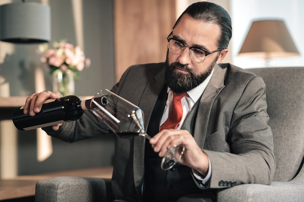 Czerwone wino. Mężczyzna w okularach i czerwonym krawacie wlewa do kieliszka czerwone wino po długim męczącym dniu