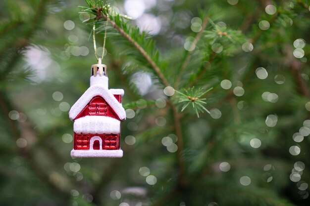 Czerwony świąteczny dom wisi na zielonej gałęzi choinki ze srebrnymi światłami bokeh.