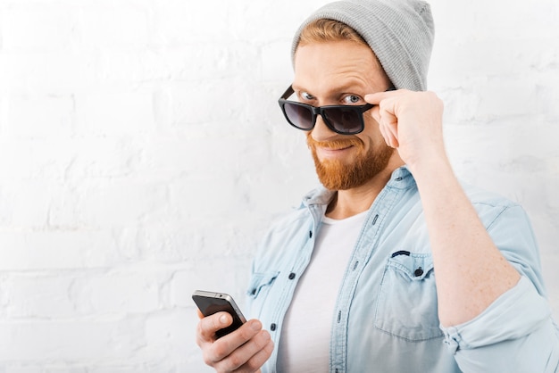 Cześć! Pewny siebie młody brodaty mężczyzna dopasowujący okulary i trzymający telefon komórkowy, stojąc przy ścianie z cegły