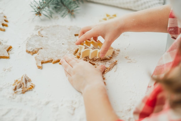 Częściowo zamazane rączki dziewczynki wycięły choinkowe pierniczki z rozwałkowanego ciasta w białej kuchni