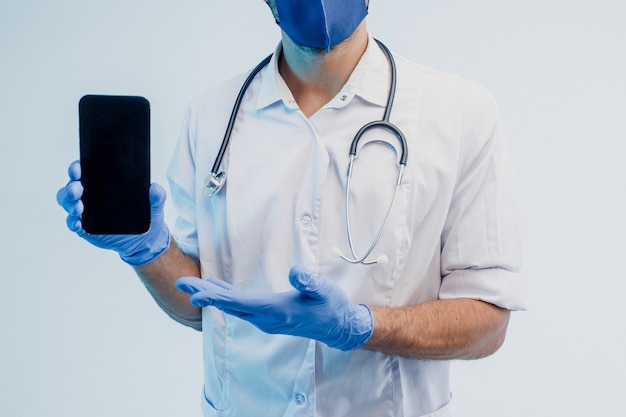Częściowy obraz europejskiego męskiego lekarza pokazując telefon komórkowy. Mężczyzna ubrany w biały fartuch, maskę ochronną i lateksowe rękawiczki ze stetoskopem. Na białym tle na szarym tle z turkusowym światłem. Sesja studyjna