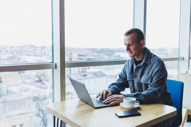 Człowiek odnoszący sukcesy szczęśliwy w biznesie siedzi przy stoliku w kawiarni, trzymając filiżankę kawy i używając laptopa