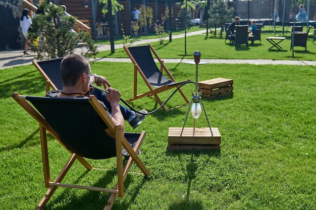 Człowiek palenia shesha, fajki wodne w przyrodzie w ciepły słoneczny dzień. Szisza wschodnia Palenie, aktywny wypoczynek na świeżym powietrzu. Leżaki na zielonej trawie. Miejsce na długi wypoczynek,