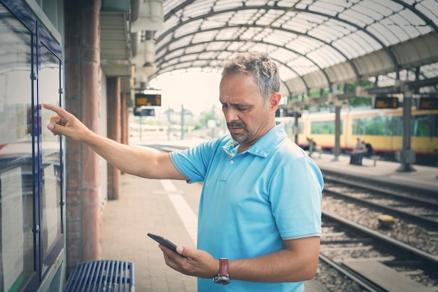 Człowiek ze smartfonem stojący na peronie przed rozkładem jazdy