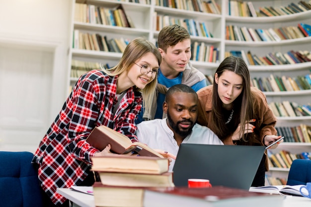 Czterech radosnych wielorasowych studentów w bibliotece, którzy razem studiują i przygotowują się do egzaminów, używając laptopa do wyszukiwania informacji w Internecie