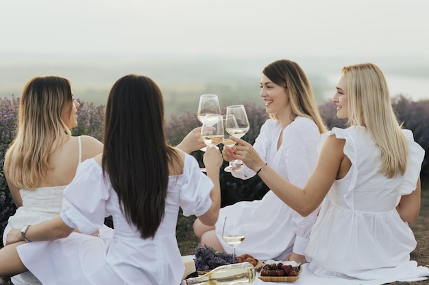 Cztery kobiety w białych szatach siedzą na kocu piknikowym i wznoszą toast kieliszkami do wina.