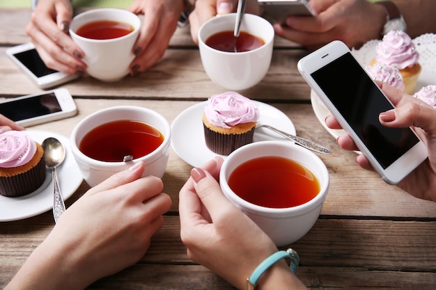 Cztery ręce ze smartfonami trzymającymi kubki z herbatą na drewnianym stole w tle