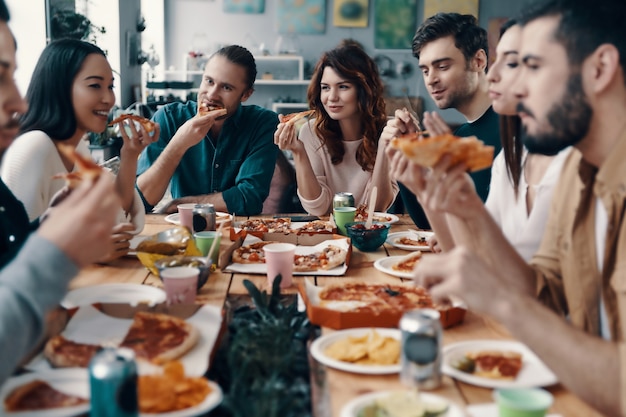 Czuć się głodnym. Grupa młodych ludzi w strojach codziennych jedzących pizzę i uśmiechających się podczas kolacji w pomieszczeniu