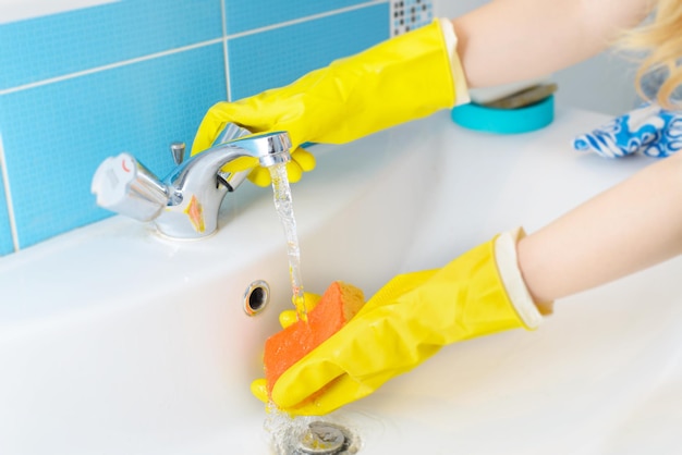 Czyszczenie czyszczenie umywalki łazienkowej i kranu z detergentem w żółtych gumowych rękawiczkach z pomarańczową gąbką
