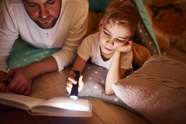 Czytanie książki i używanie latarki Ojciec i jego młode spędzają razem czas w pomieszczeniu w pobliżu namiotu