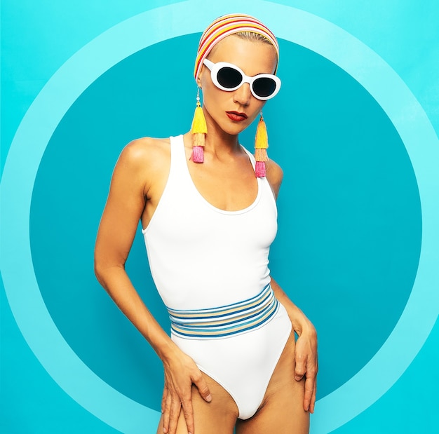 Dama w stylowych akcesoriach plażowych. Okulary przeciwsłoneczne, kolczyki, biały strój kąpielowy. Modny wygląd plaży