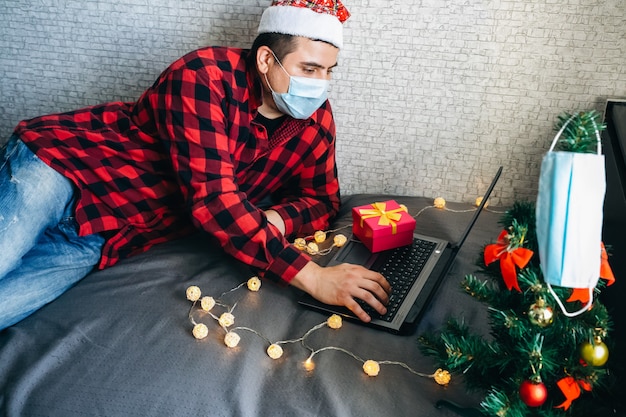 Dawanie prezentów świątecznych. Mężczyzna w masce medycznej udostępnianie prezentów w pudełku za pośrednictwem połączeń wideo. Wakacje w odosobnieniu. Nowy rok drzewo i światła z dekoracjami. Rodzina i przyjaciele świętują online.