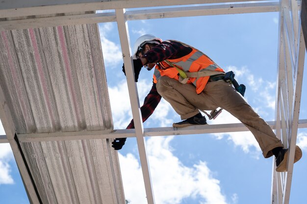 Zdjęcie dekarz pracujący w ochronnych rękawicach roboczych, pracownik budowlany noszący szelki bezpieczeństwa pracujący na wysokim poziomie na placu budowy, montuje nowy dach, metalowy dach.