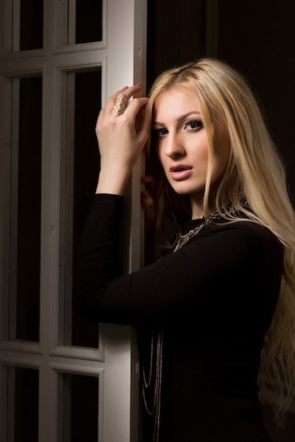Delikatna blondynka z długimi bujnymi włosami pozuje przy białych drzwiach z cieniem na twarzy