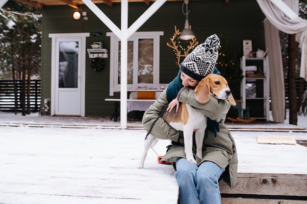 Delikatna kobieta siedzi na zaśnieżonym tarasie przed domem, przytula swojego psa rasy beagle stojącego na jej kolanach i pochylając się do niej.