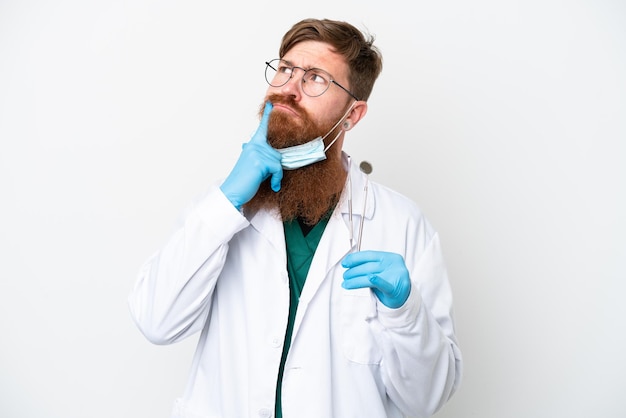 Dentysta czerwonawy mężczyzna trzyma narzędzia odizolowane na białym tle mając wątpliwości patrząc w górę