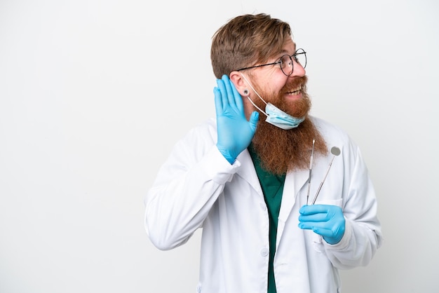 Dentysta czerwonawy mężczyzna trzymający narzędzia odizolowane na białym tle, słuchając czegoś, kładąc rękę na uchu