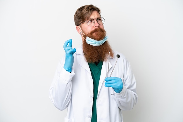 Dentysta czerwonawy mężczyzna trzymający narzędzia odizolowane na białym tle z palcami skrzyżowanymi i życzącymi wszystkiego najlepszego