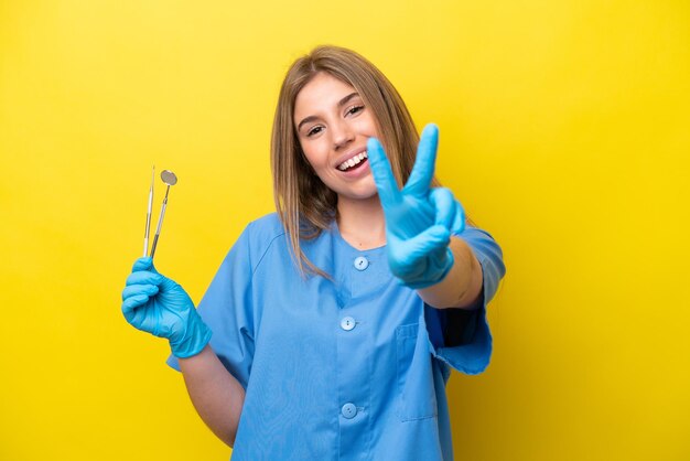 Dentysta kaukaska kobieta trzyma narzędzia odizolowane na żółtym tle, uśmiechając się i pokazując znak zwycięstwa