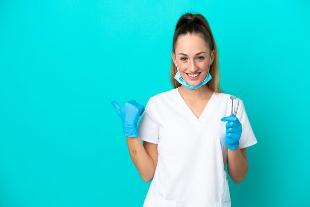 Dentysta kaukaska kobieta trzymająca narzędzia izolowane na niebieskim tle wskazujące na bok, aby zaprezentować produkt