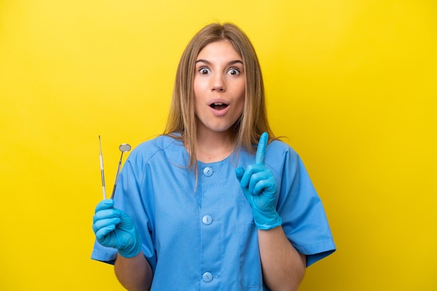 Dentysta kaukaska kobieta trzymająca narzędzia izolowane na żółtym tle, zamierzająca zrealizować rozwiązanie, podnosząc palec w górę